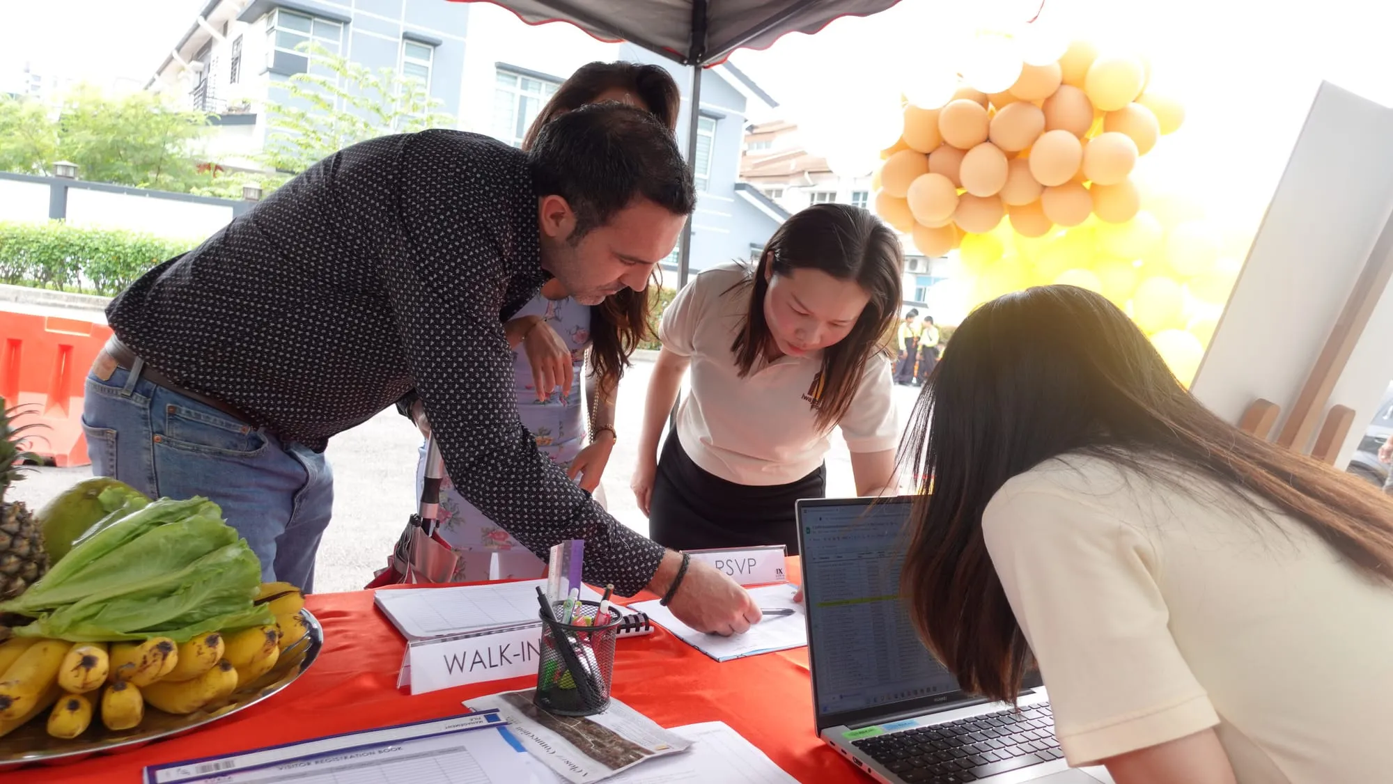 Guests signing in at the walk-in desk, the fruit tray and the balloon bunches beside them.