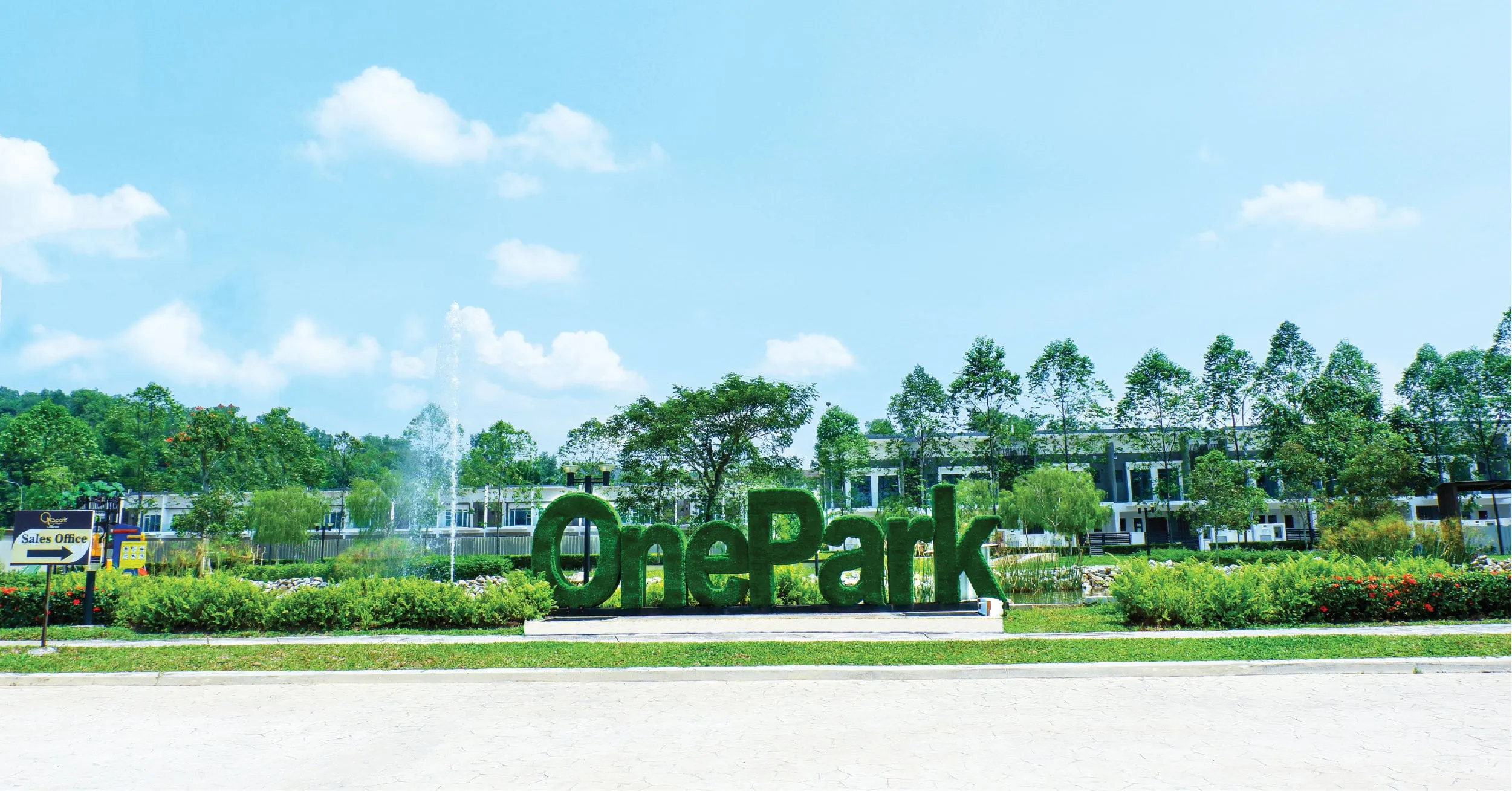 Hedge letters spelling “OnePark” on a low kerb in the landscaped grounds of the completed OnePark development, Taman Semenyih Idaman, with a fountain jet, a Sales Office signpost and rows of terrace homes behind.